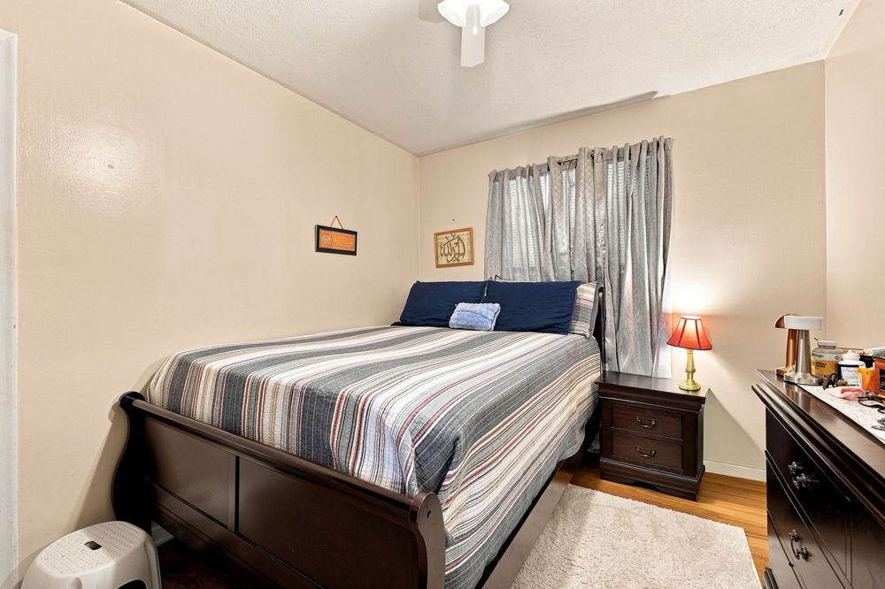 Cozy bedroom with a queen-sized bed, striped bedding, hardwood floors, and curtained window with warm lighting.