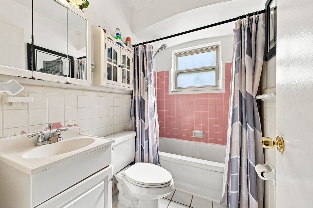 Vintage bathroom with pink tile tub surround, white toilet, pedestal sink, mirrored cabinet, and striped shower curtain.