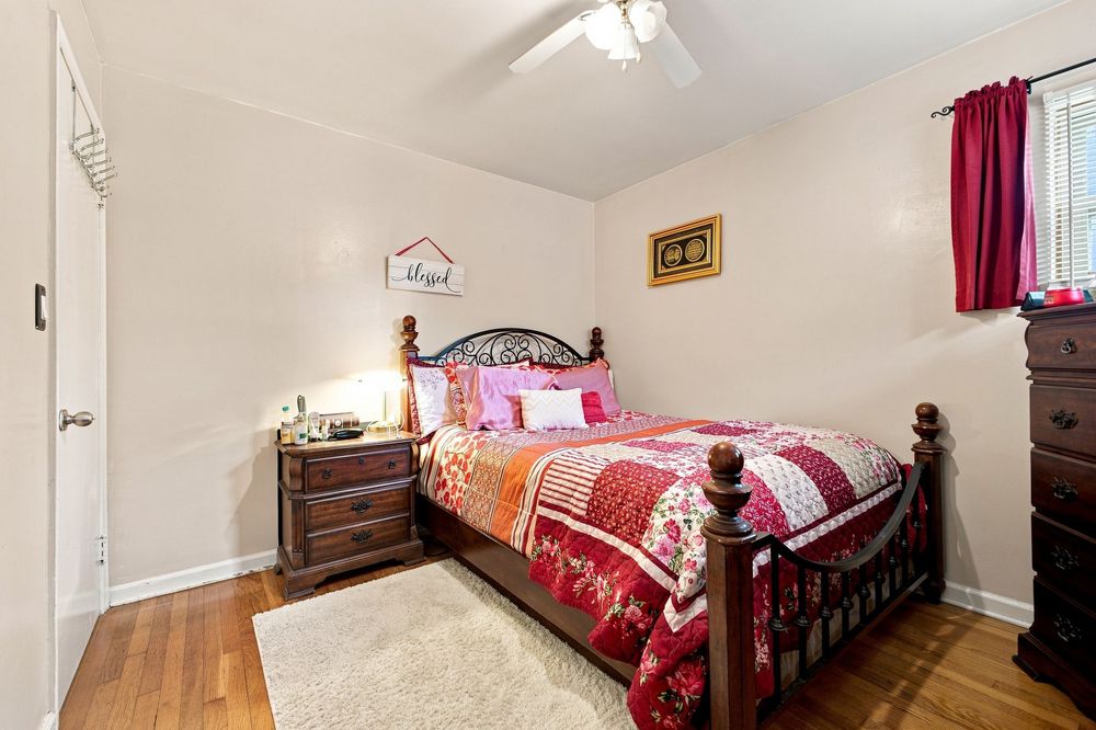 Cozy bedroom with a queen-sized wooden bed, red patchwork quilt, hardwood floors, ceiling fan, and dark wood dresser.
