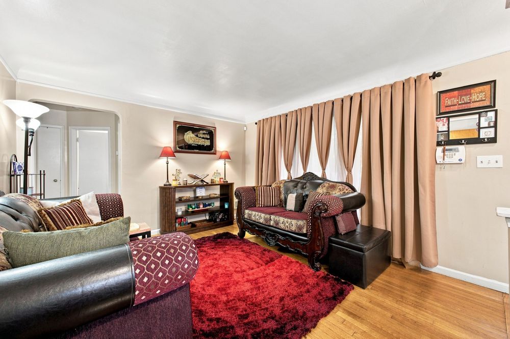 Cozy living room with hardwood floors, red shag rug, leather sofa, and warm beige curtains framing the windows.