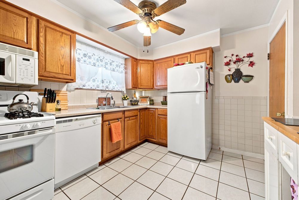 Bright kitchen with oak cabinets, white appliances, tile flooring, and a ceiling fan above a window with natural light.