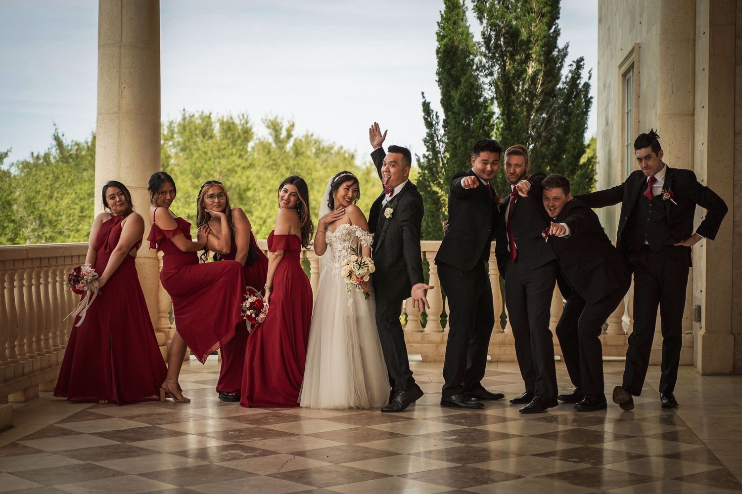 Wedding party poses on grand pillared balcony with bridesmaids in burgundy dresses and groomsmen in black suits.
