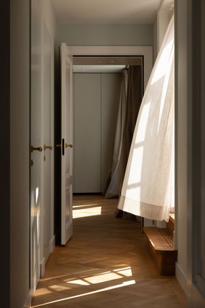 Sunlit hallway with herringbone parquet floors, white doors, and sheer linen curtains billowing in natural light.