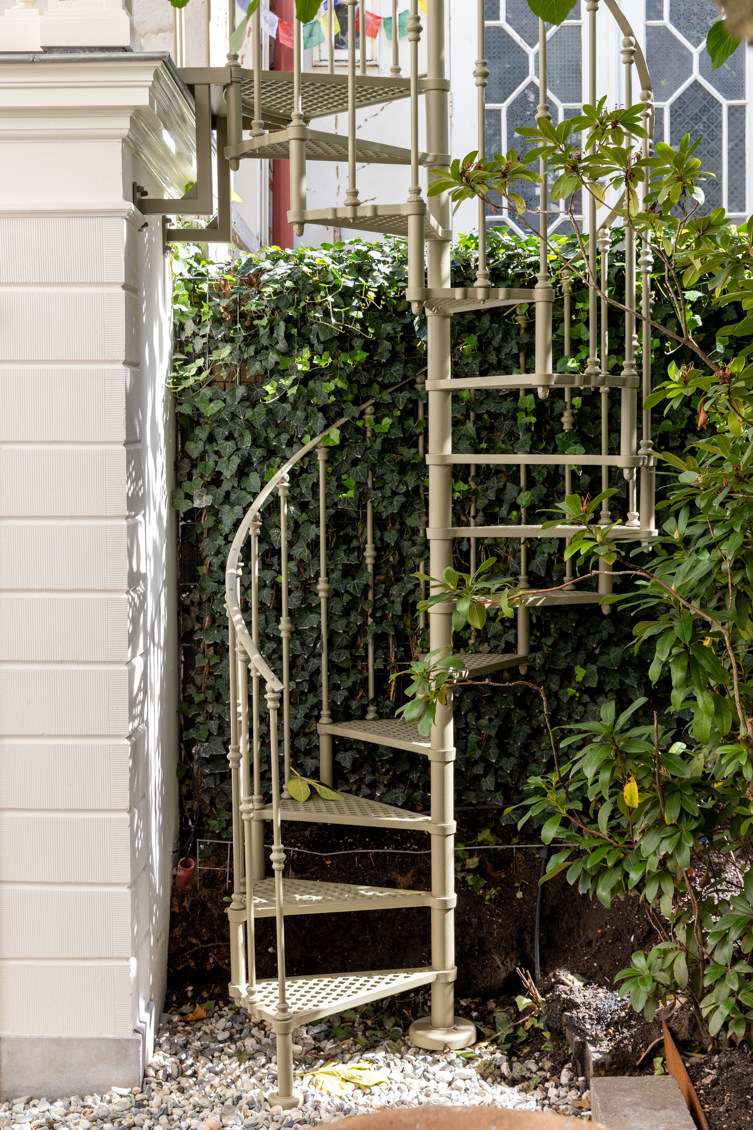 Outdoor metal spiral staircase with ornate railings surrounded by lush green ivy and plants against a white building wall.