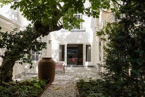 Elegant white villa courtyard with lush greenery, stone pathway, and large terracotta urn surrounded by mature trees.