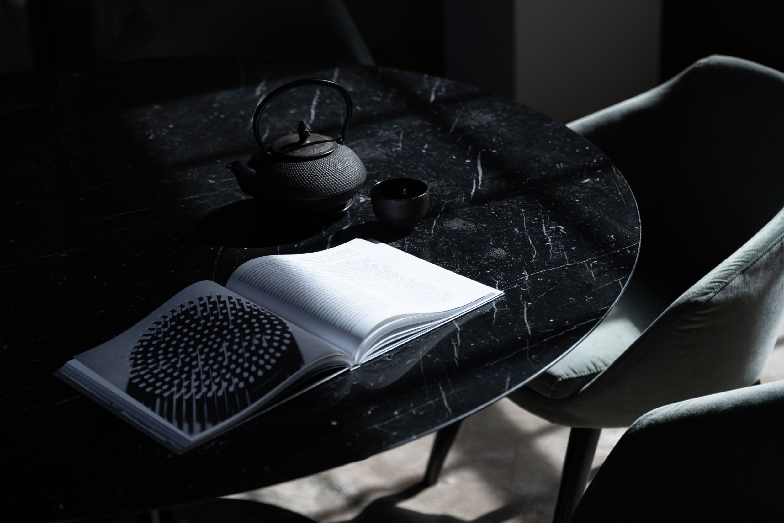 Dark marble table with open book, black cast iron teapot, and decorative mat beside a modern chair in moody lighting.
