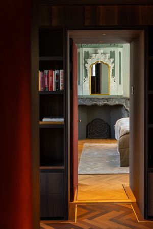 Elegant bedroom viewed through doorway with ornate gold mirror above fireplace, bookshelf with red books, and herringbone wood floors.