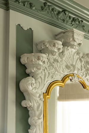 Ornate white Baroque plaster molding with gold-framed mirror against sage green wall paneling.