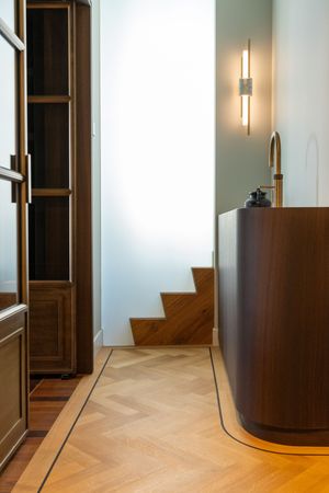 Elegant bathroom with curved walnut vanity, herringbone parquet floor, wooden bookcase, and modern wall sconce light.