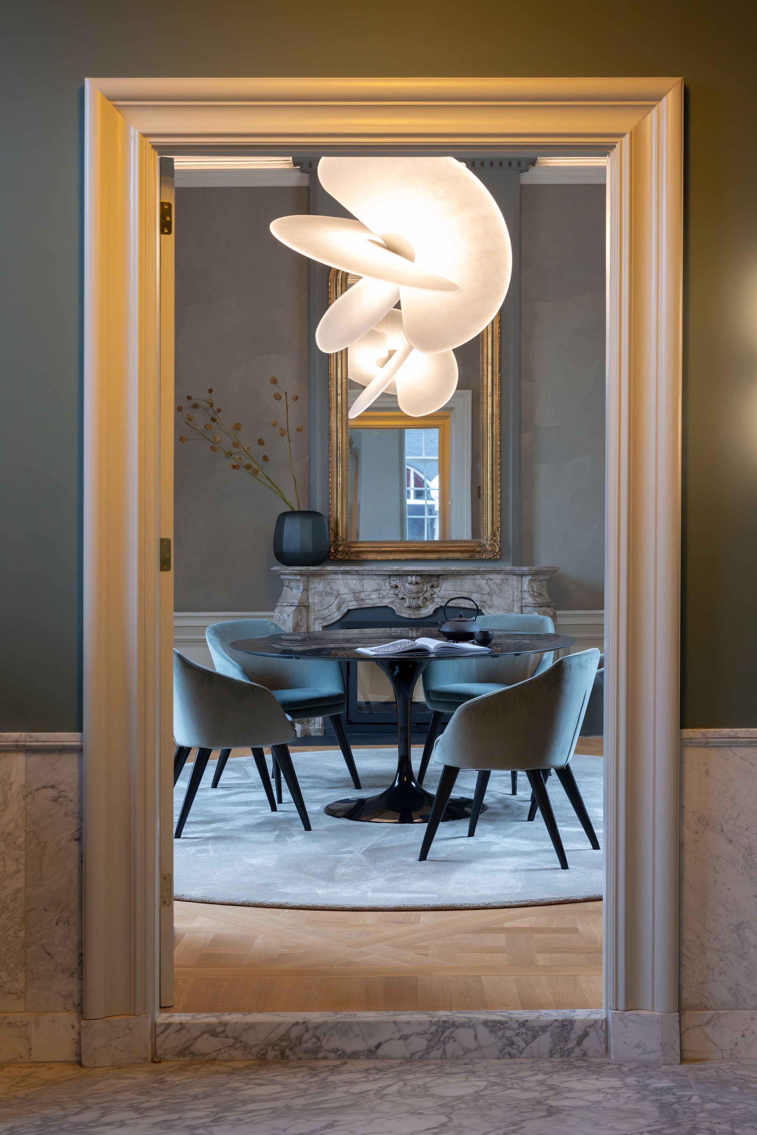 Elegant dining room viewed through doorway with modern pendant light, round black table, gray chairs, and ornate gold mirror.