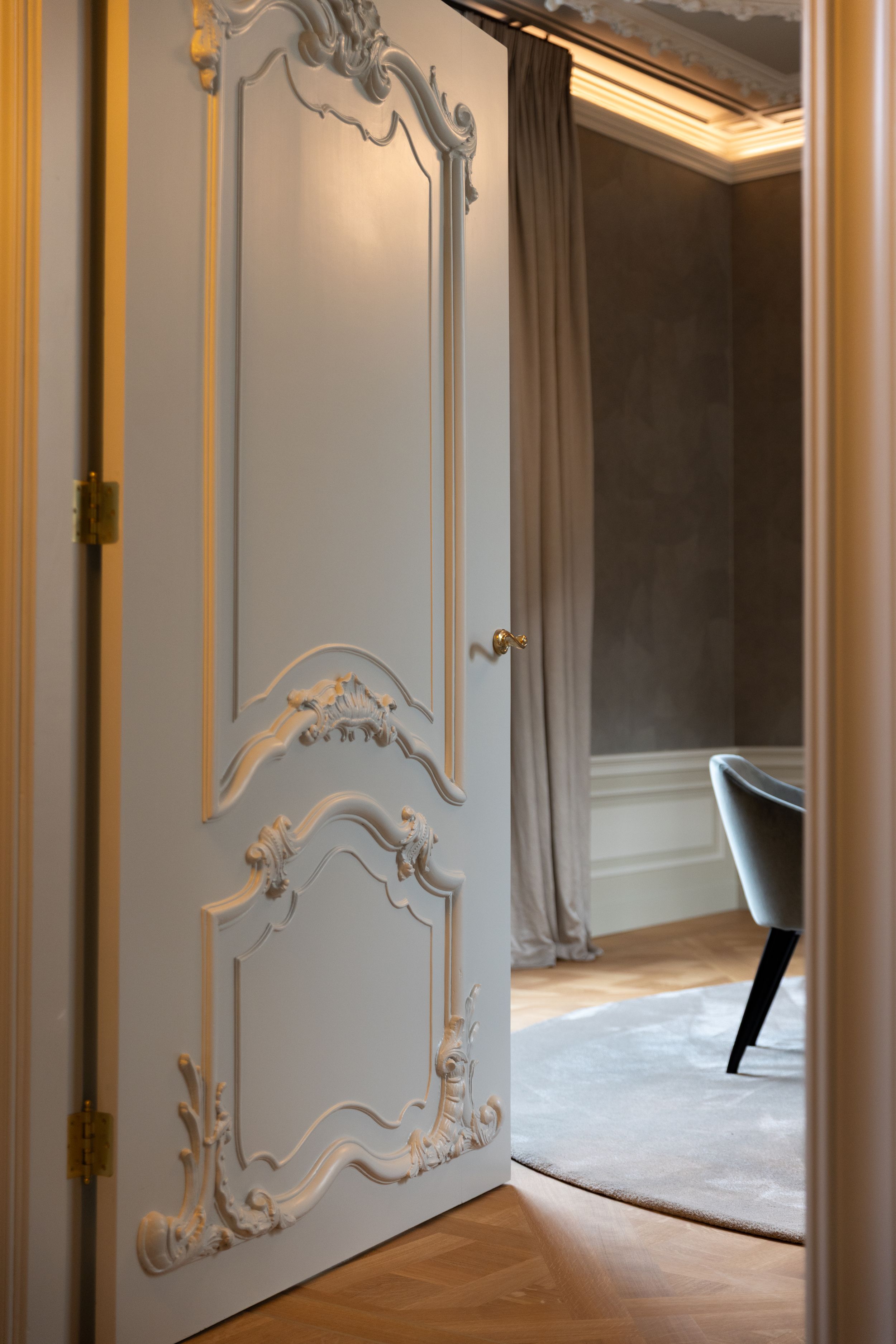 Elegant French door with ornate white rococo moldings opens to a sophisticated room with gray walls and herringbone floors.