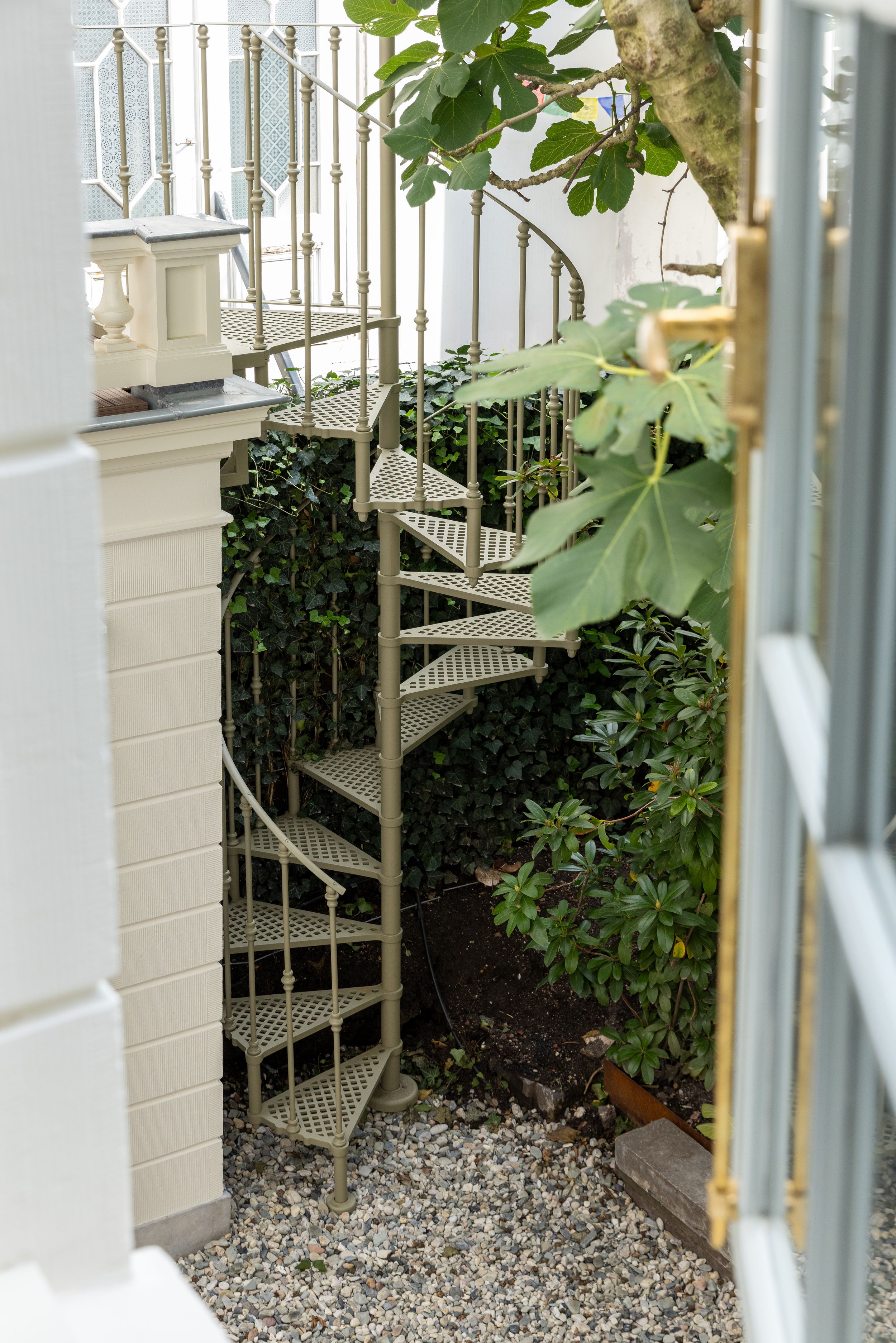 Elegant outdoor spiral staircase with ornate metal railings surrounded by lush green garden plants and shrubs.