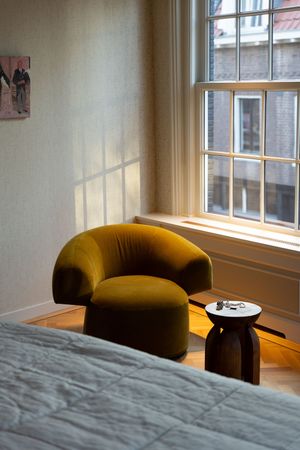 A mustard yellow velvet armchair beside a small dark wood side table near a sunlit window in a cozy bedroom.