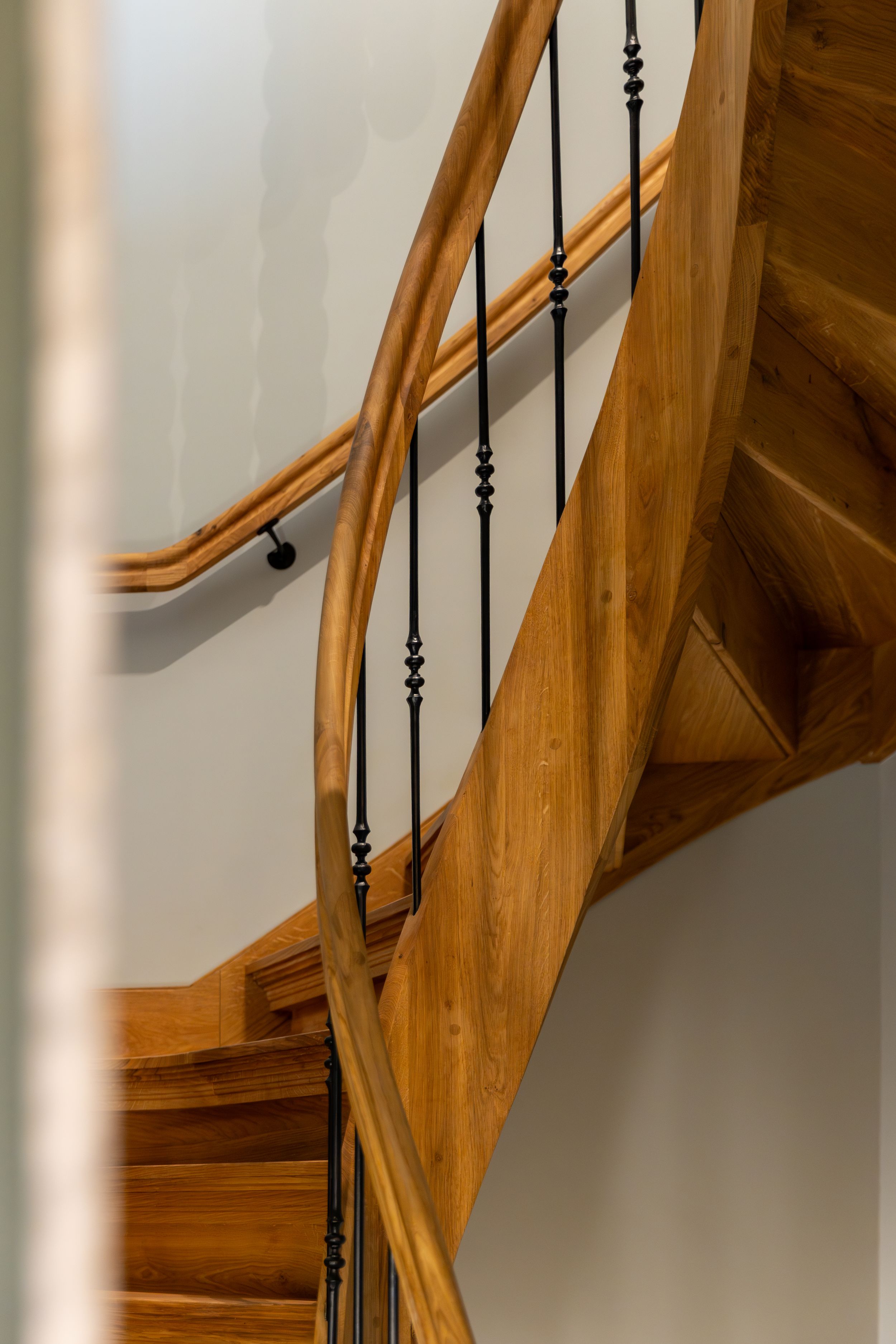 Curved wooden spiral staircase with ornate black iron balusters and smooth oak handrail in classic interior design.