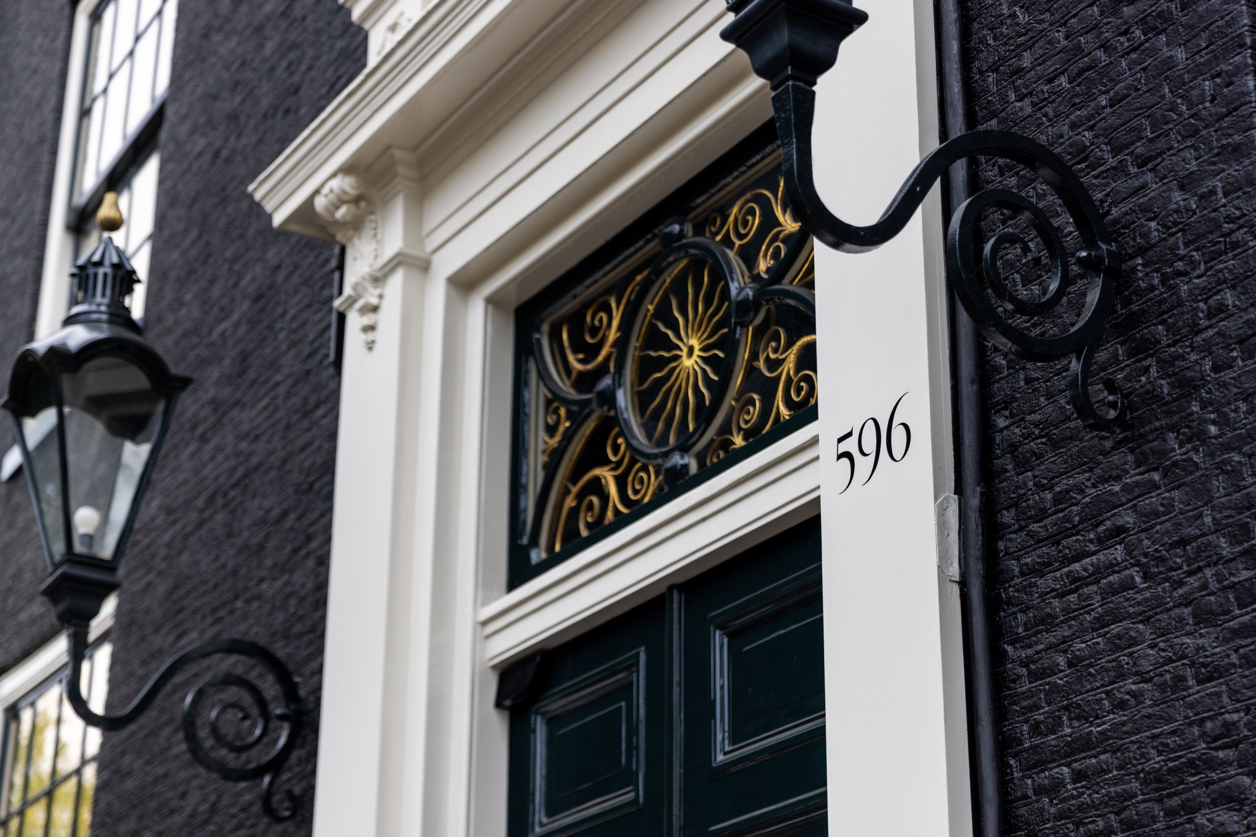 Elegant black door numbered 596 with ornate gold fanlight and wrought iron lamp on a classic Amsterdam canal house.