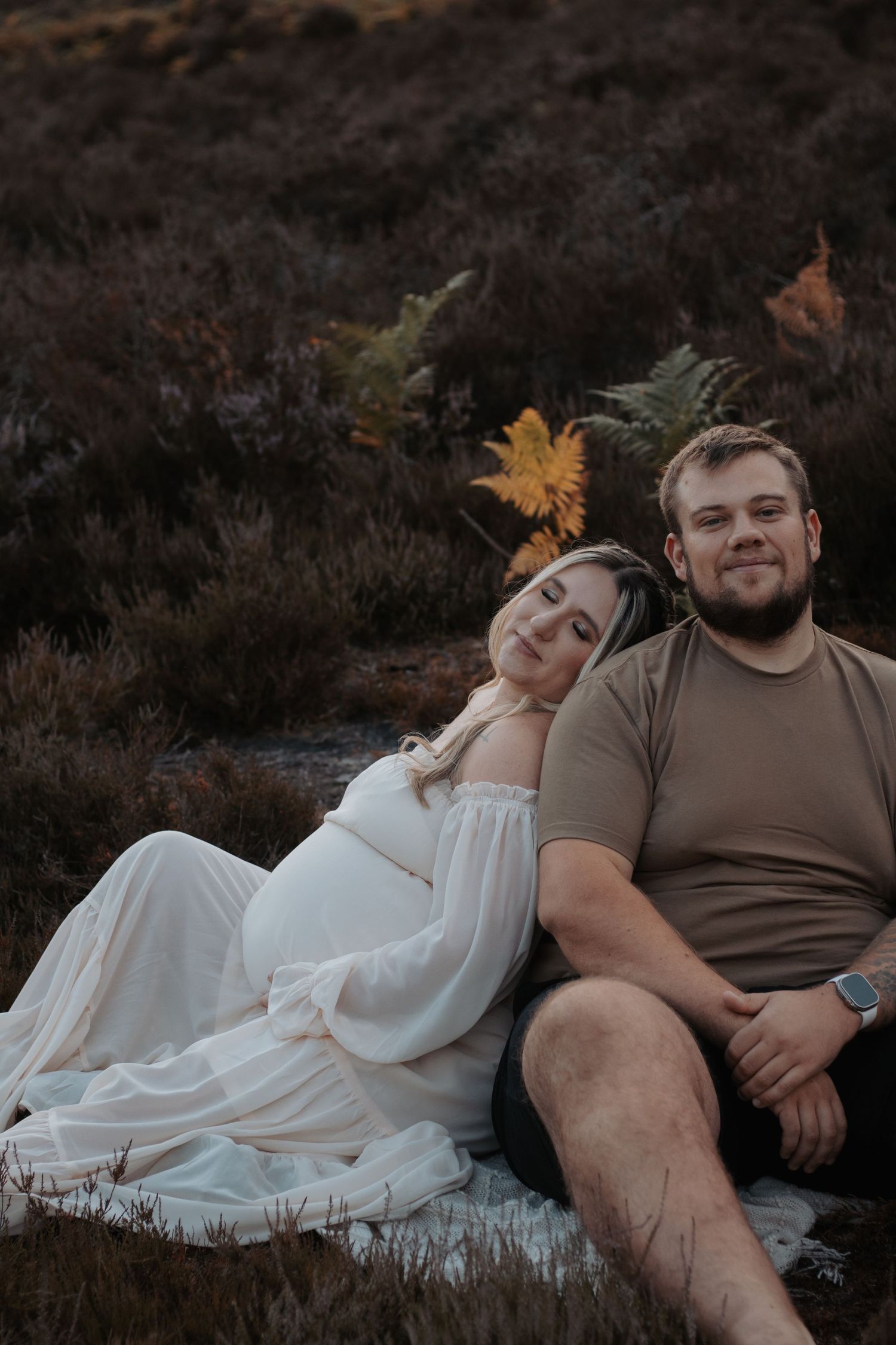 Pregnant woman in white dress leans on partner sitting outdoors in autumn heathland for maternity photo.