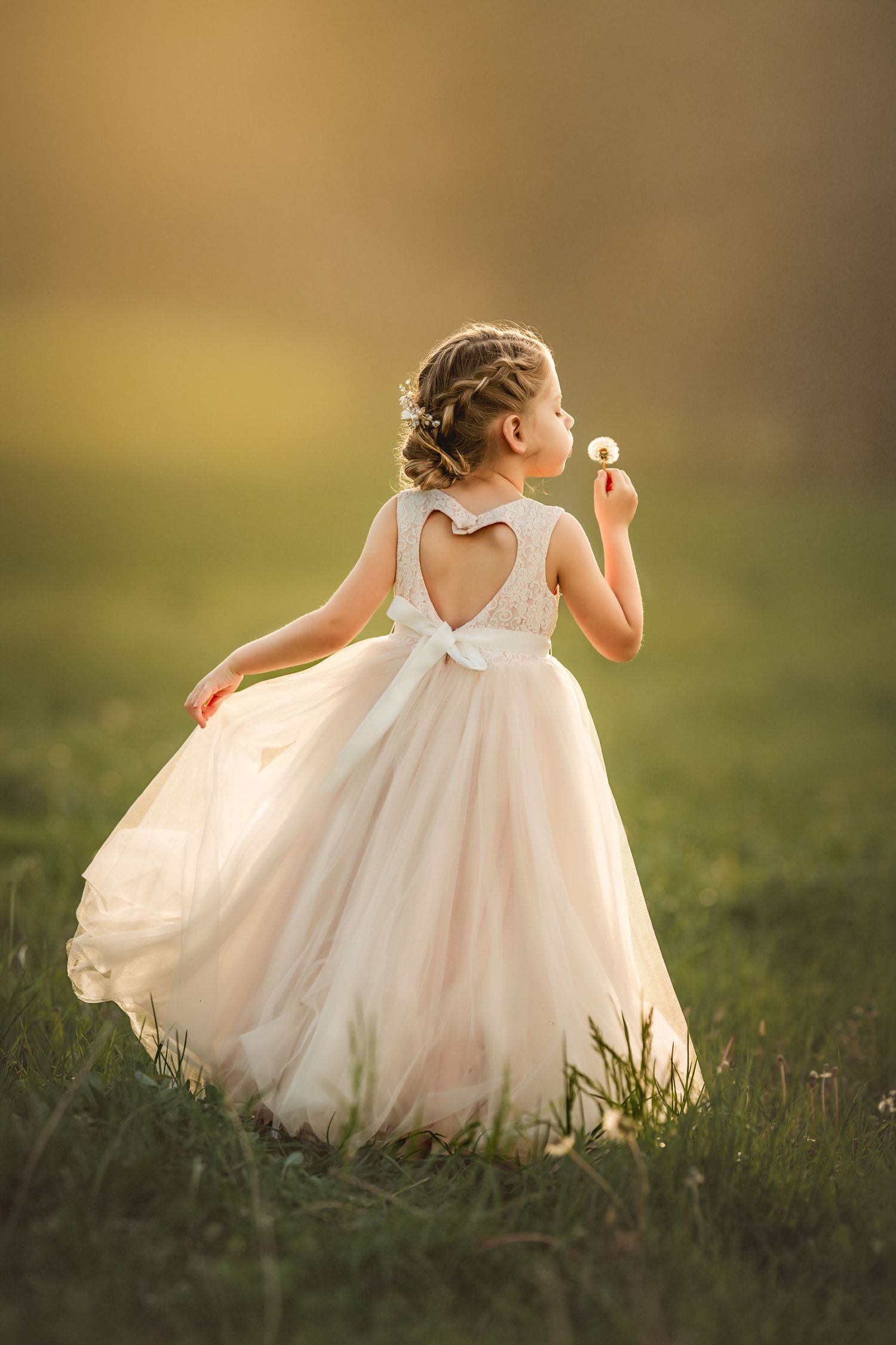 Young girl in elegant blush tulle gown blowing dandelion in sunlit meadow, heart cutout back detail.