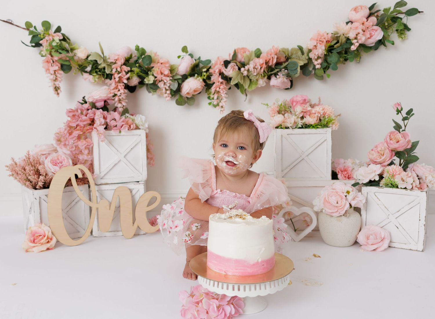 Baby girl in pink outfit celebrates first birthday with smash cake surrounded by pink floral decorations and One sign.