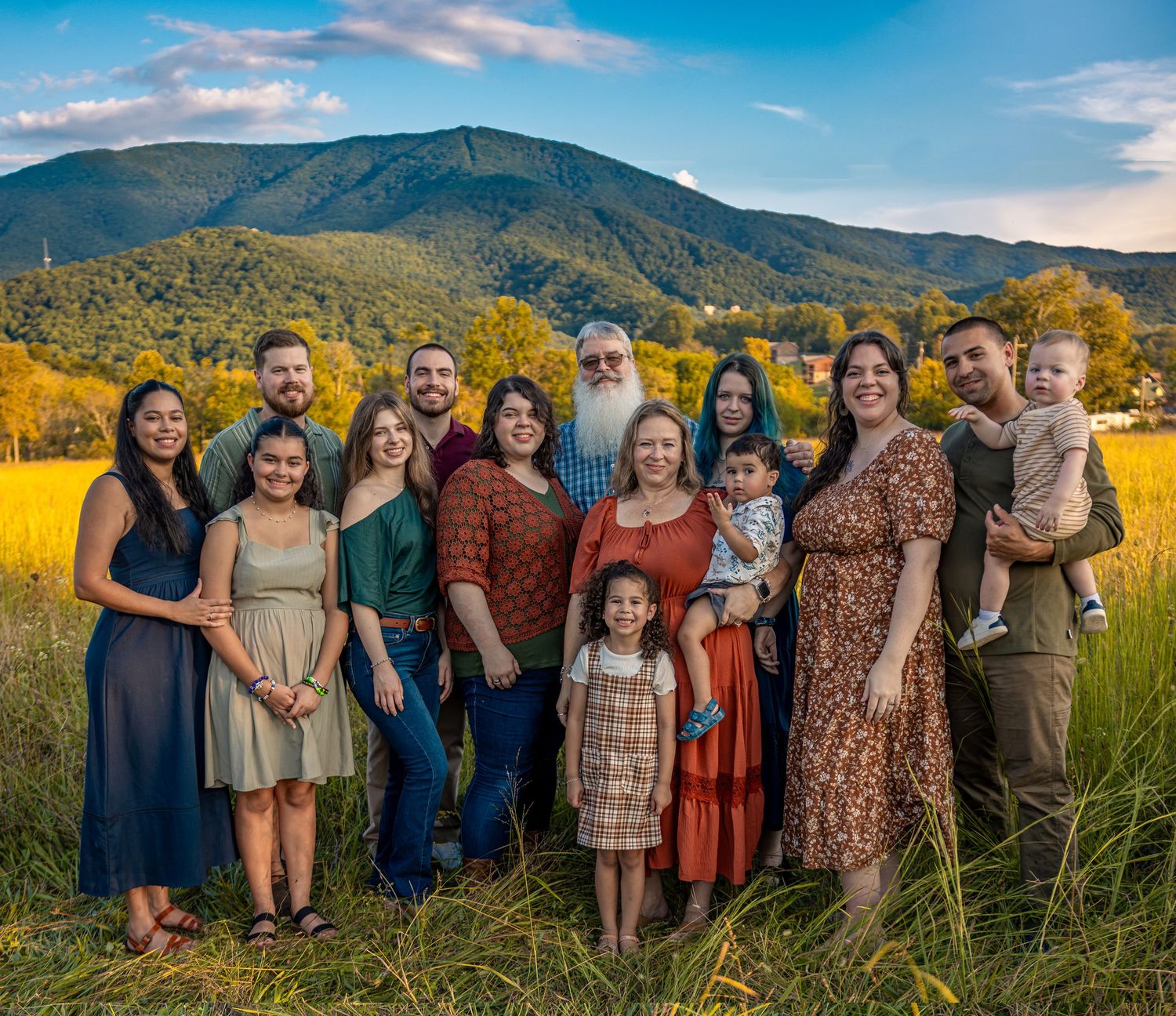 Large family group smiling outdoors with scenic mountain and blue sky backdrop on a sunny summer day.