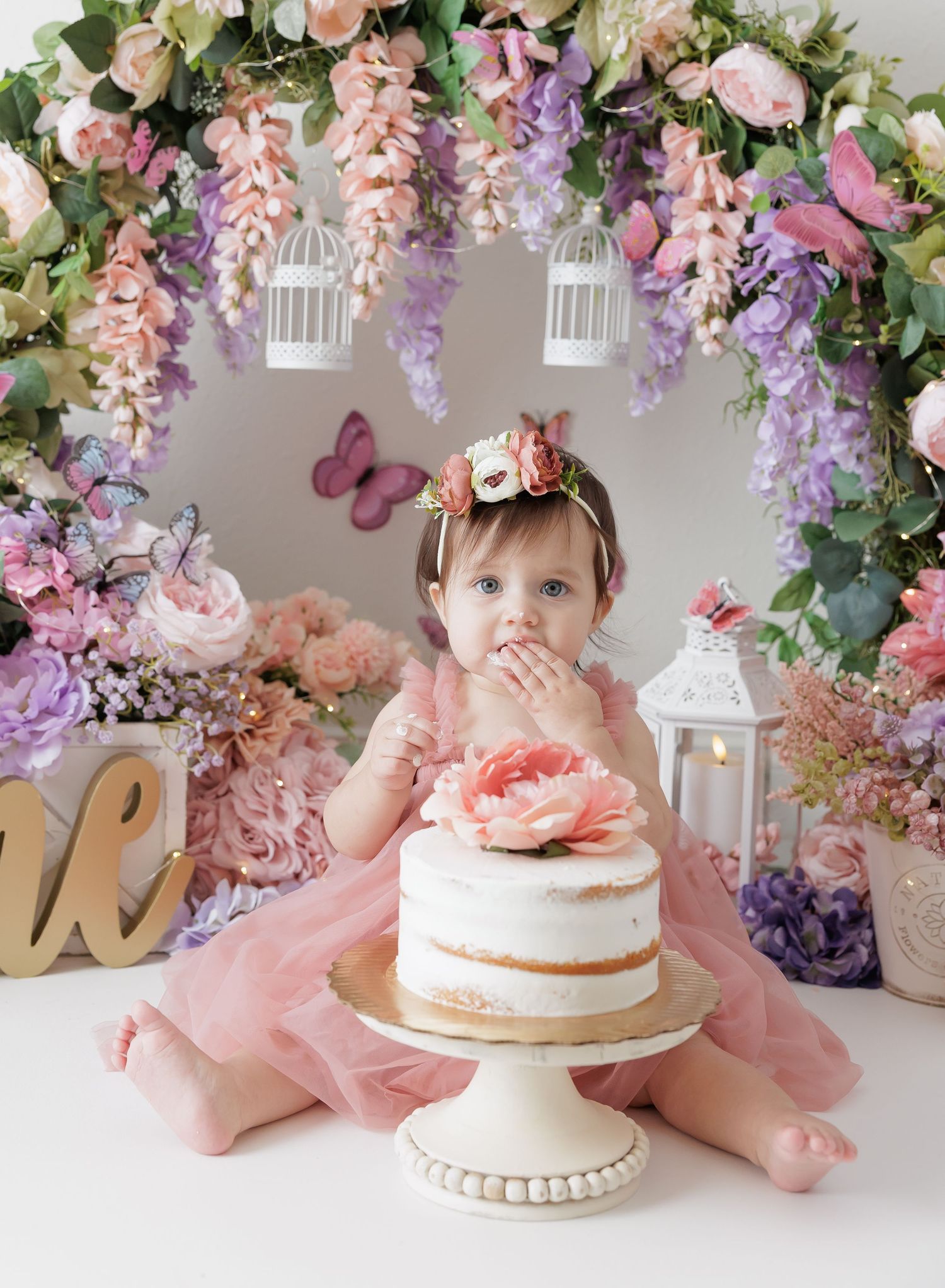 Baby girl in pink tutu sits before floral arch for first birthday cake smash photo session.