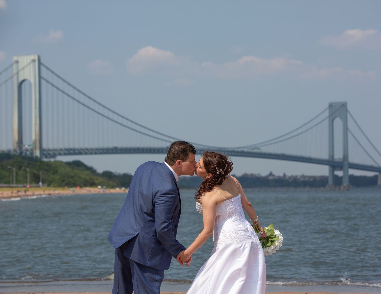 Bride and groom kissing on beach with Verrazzano-Narrows Bridge in background on sunny wedding day.