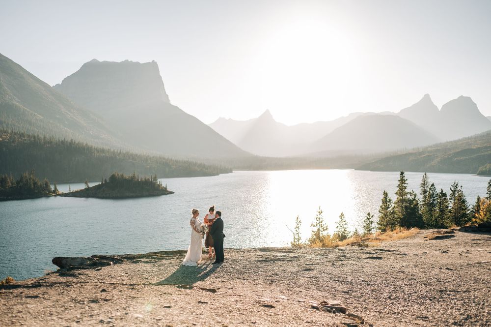 Couple exchanges vows at stunning mountain lake overlook with dramatic peaks and golden sunlight in background.