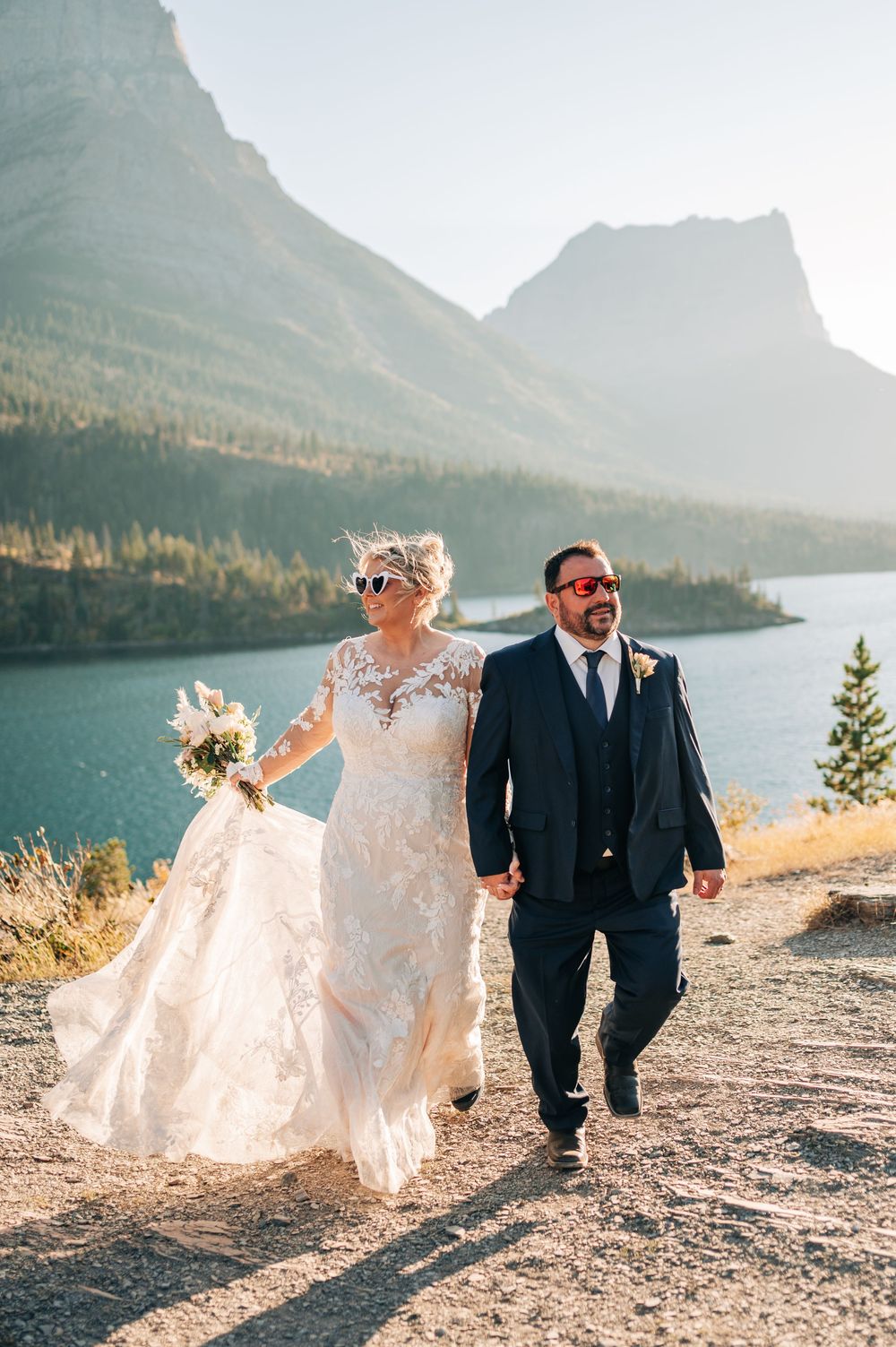 Bride and groom walk hand-in-hand along a scenic mountain lake path, bride in lace gown, groom in navy suit.