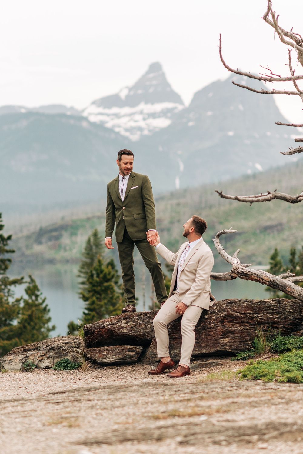 Two grooms in suits hold hands on rocky mountain overlook with snow-capped peaks and lake backdrop.