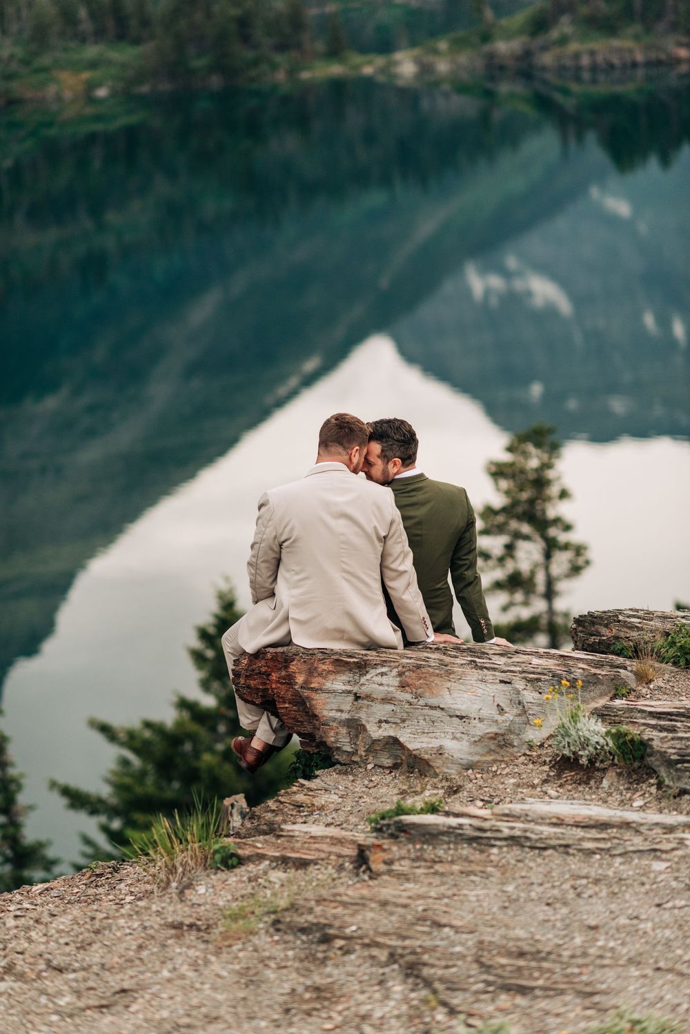 Two grooms sit on a rocky cliff edge sharing a romantic moment overlooking a serene mountain lake reflection.