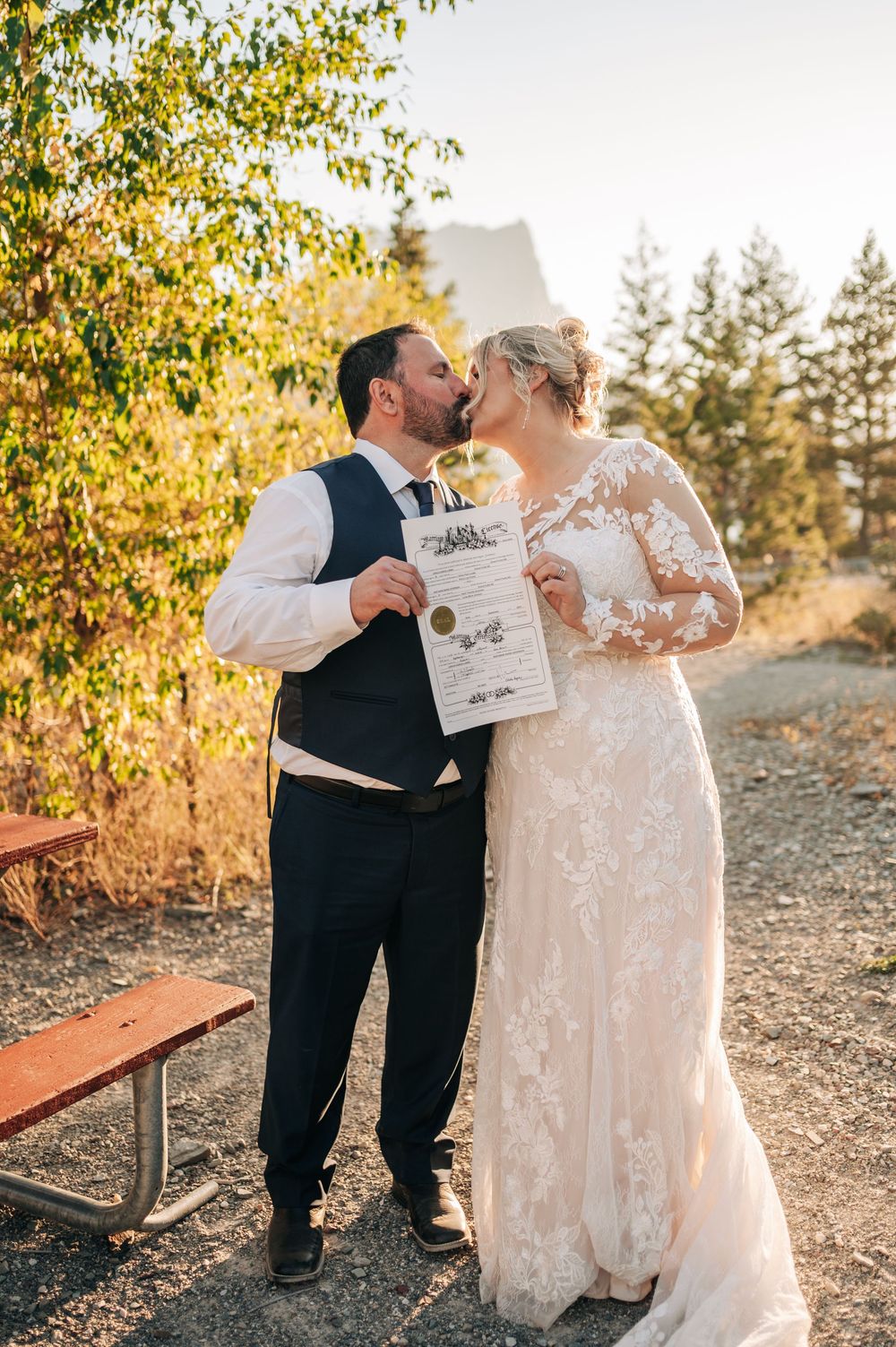 Newlywed couple kissing while holding marriage certificate outdoors in autumn setting, bride in lace gown.