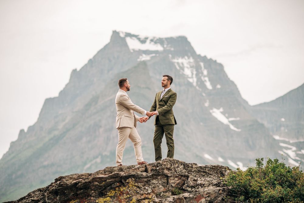 Two grooms hold hands on a rocky mountain summit with snow-capped peaks in the background during their outdoor wedding.