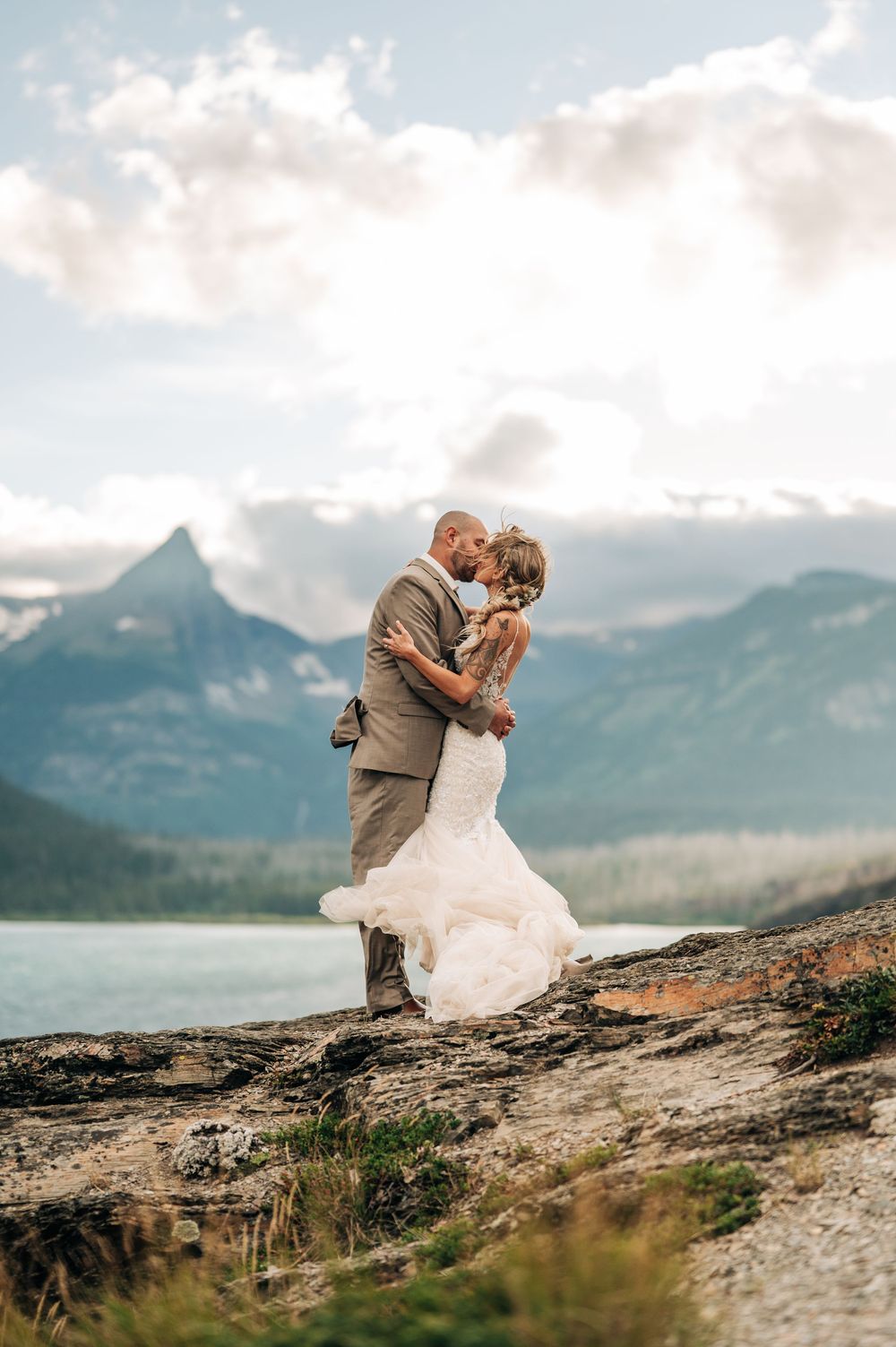 Bride and groom kissing on rocky mountain overlook with turquoise lake and peaks backdrop on wedding day.