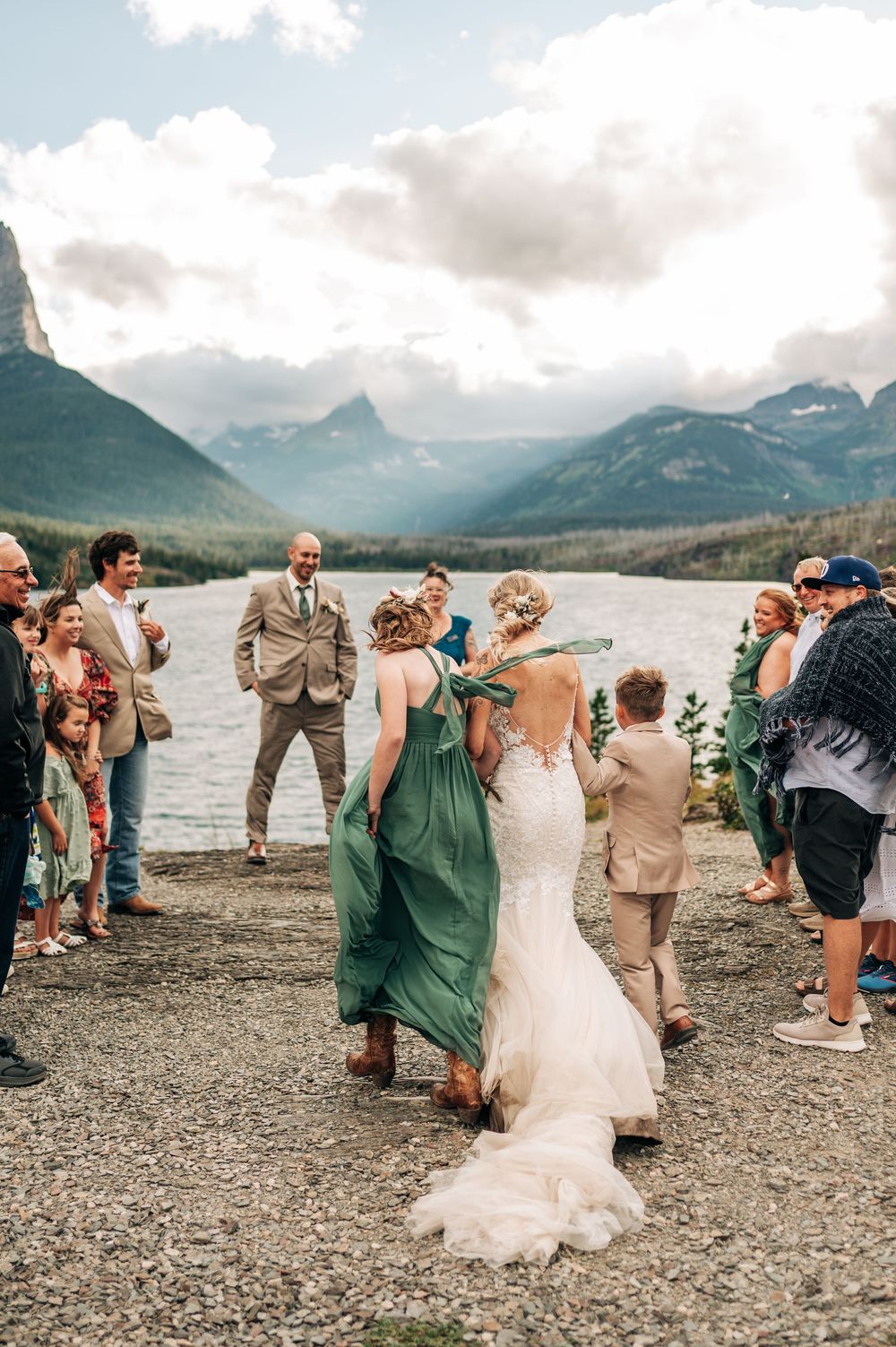 Bride in lace gown walks with bridesmaid in green dress at outdoor mountain lake wedding ceremony.
