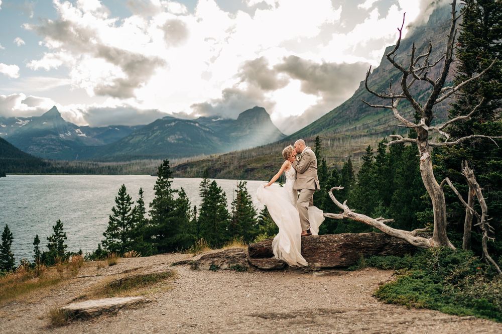 Couple in wedding attire embracing on a log with mountain lake and evergreen forest backdrop at Glacier National Park.