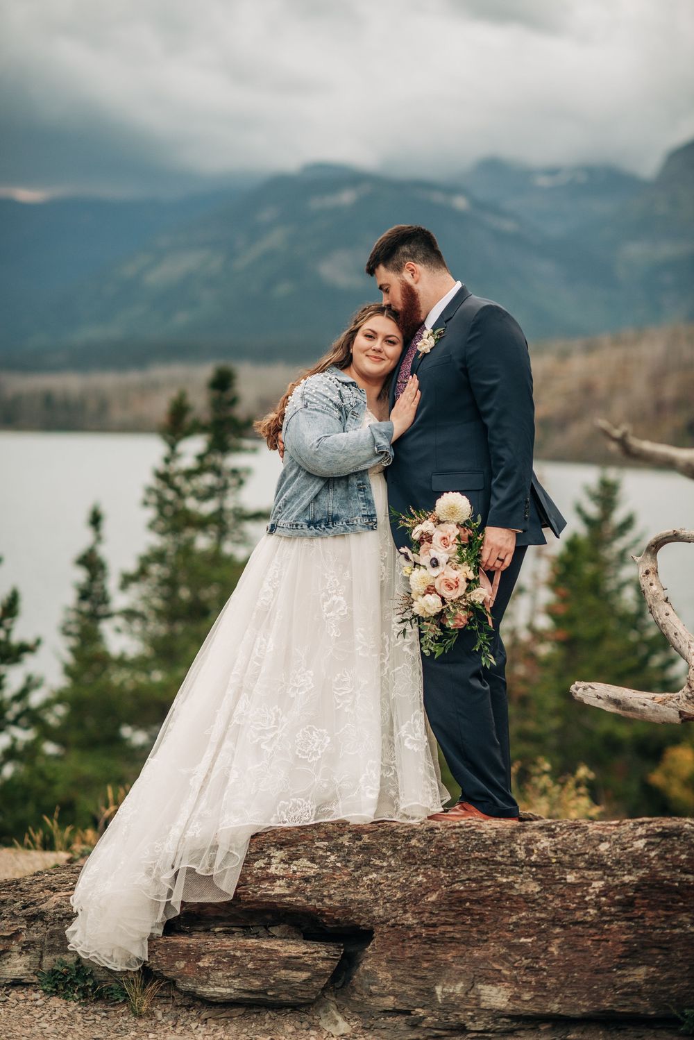 Bride in white gown and denim jacket embraces groom in navy suit outdoors with mountain lake backdrop.