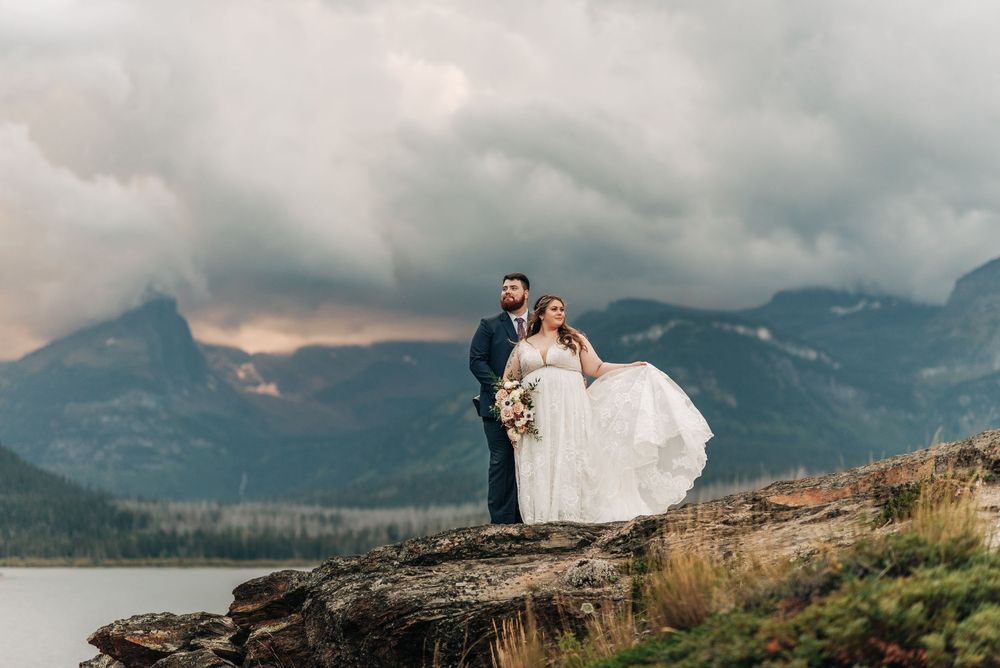 Bride and groom stand on rocky cliff with stormy mountain lake backdrop in stunning outdoor wedding portrait.