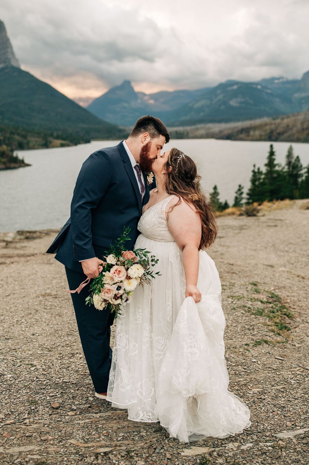 Bride and groom share a kiss at mountain lake overlook, bride holding blush bouquet, groom in navy suit.