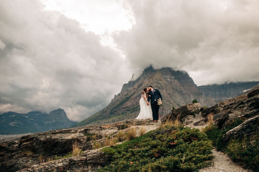 Bride and groom embracing on rocky mountain path with dramatic cloudy skies and rugged peaks in background.