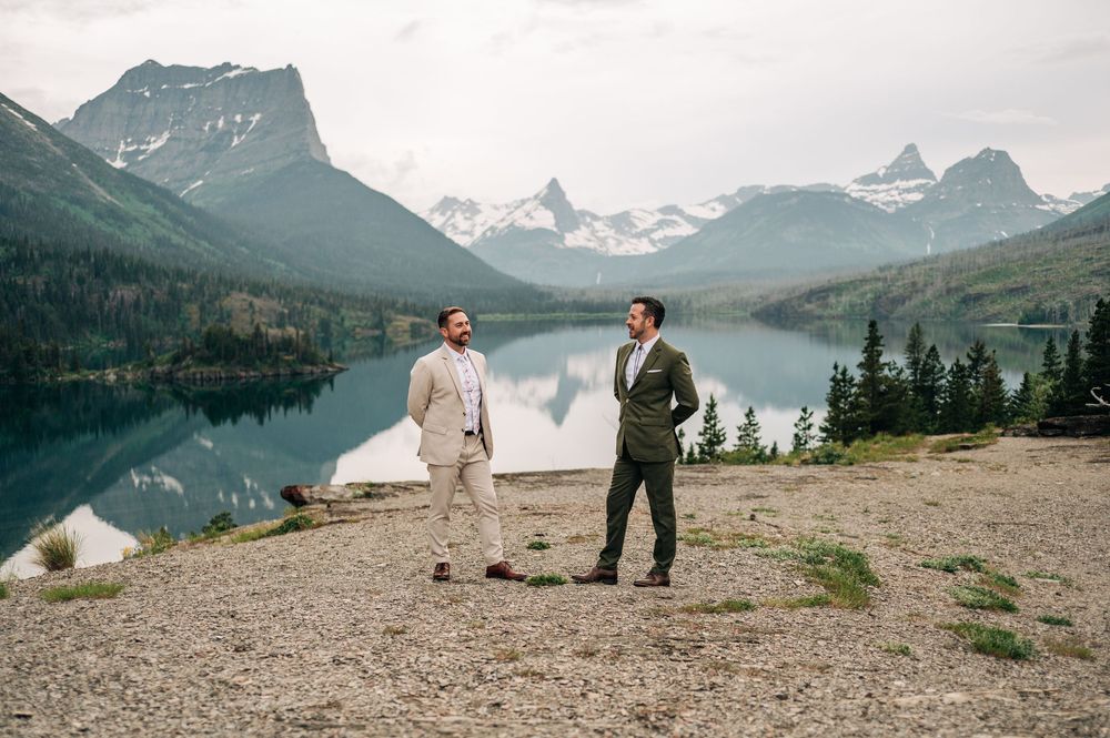 Two grooms in suits stand on rocky shore with stunning mountain lake backdrop at Glacier National Park wedding.