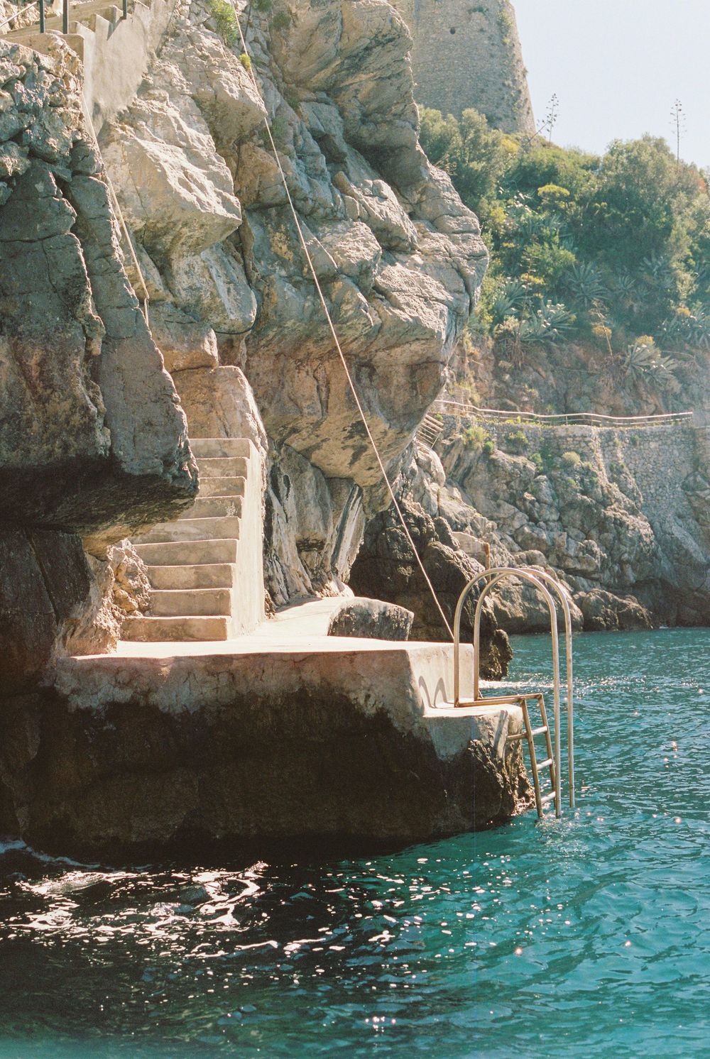 Rocky Mediterranean cliffside with stone steps, metal handrails, and a small dock platform overlooking turquoise blue water.