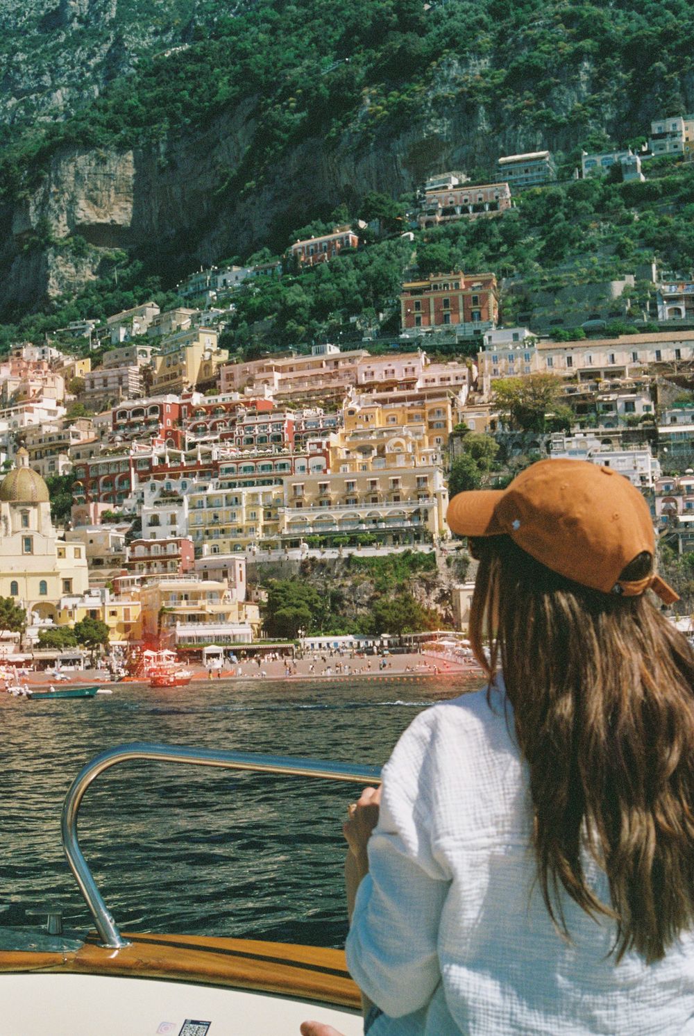 Woman in white top and brown cap views colorful Positano cliffside buildings from a boat on the Amalfi Coast.
