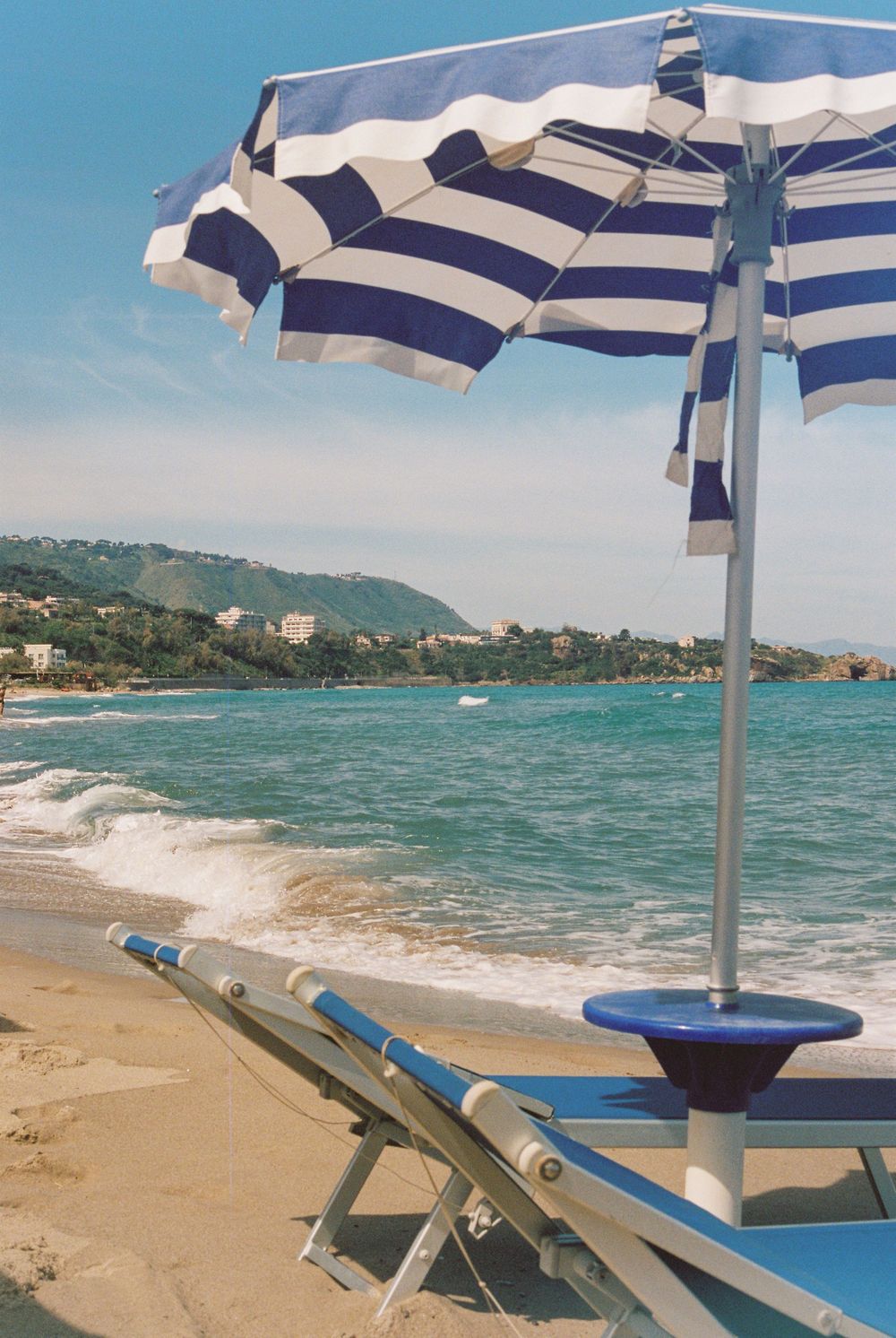 Blue and white striped beach umbrella over lounge chairs on a sunny sandy shore with turquoise ocean waves.