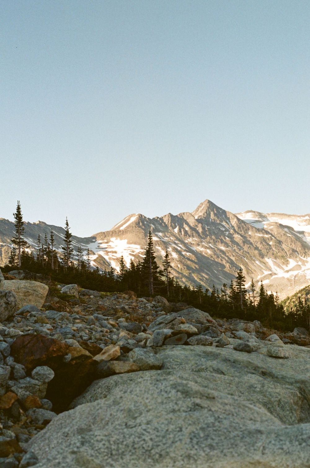 Rocky alpine landscape with snow-capped mountain peaks, evergreen trees, and golden sunlight at dusk.