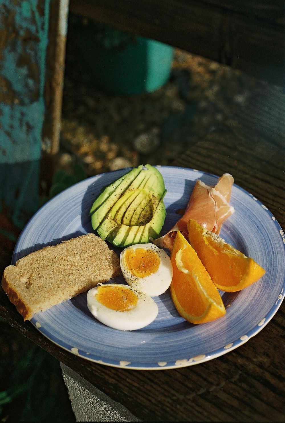 A blue plate with sliced avocado, boiled eggs, orange wedges, toast, and prosciutto served outdoors on a wooden table.