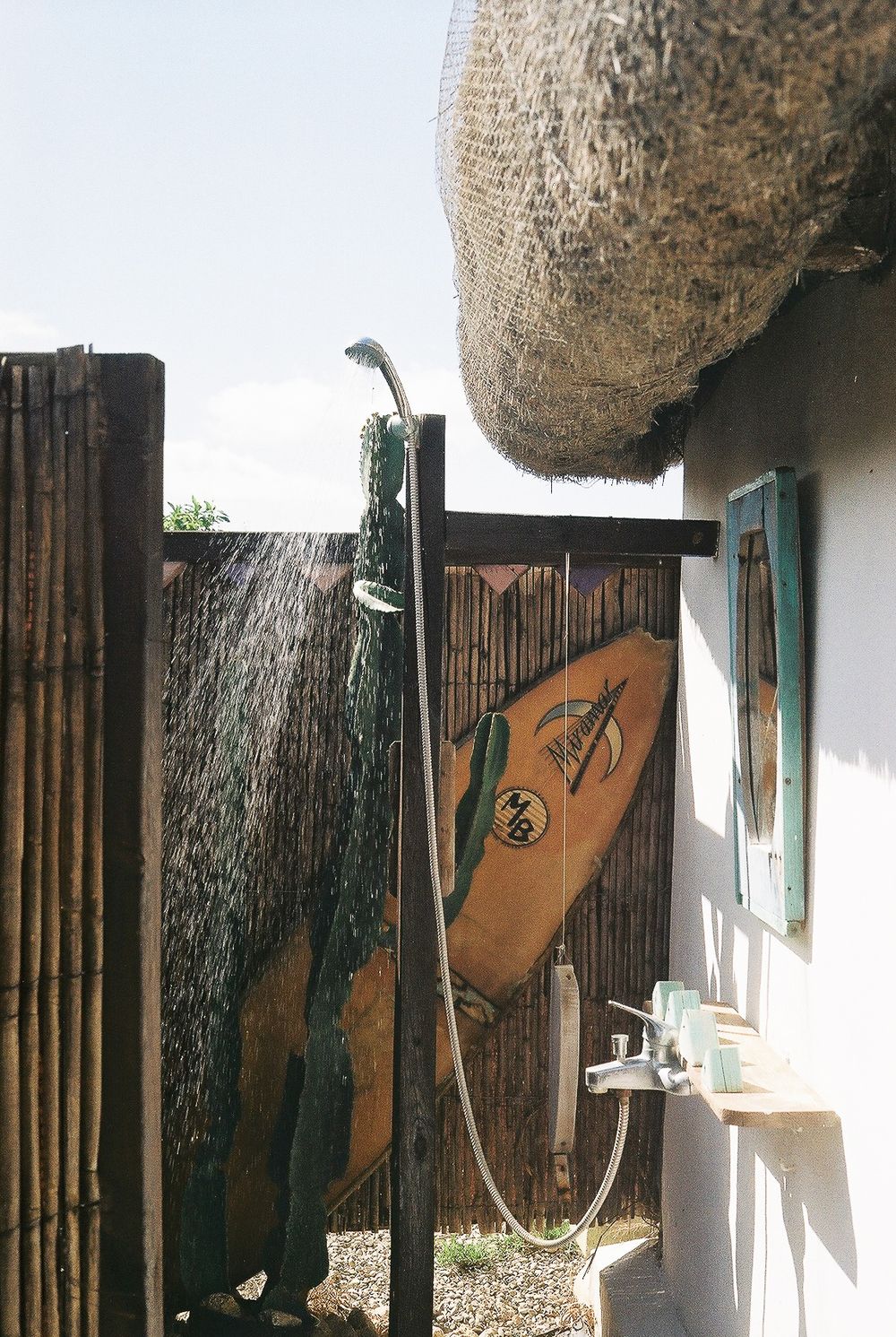 Outdoor shower with bamboo fence, surfboard leaning against wall, and thatched roof in a tropical surf retreat setting.
