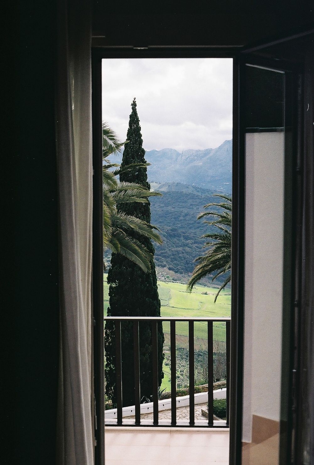 Open balcony doors revealing a scenic mountain view with tall cypress and palm trees under a cloudy sky.