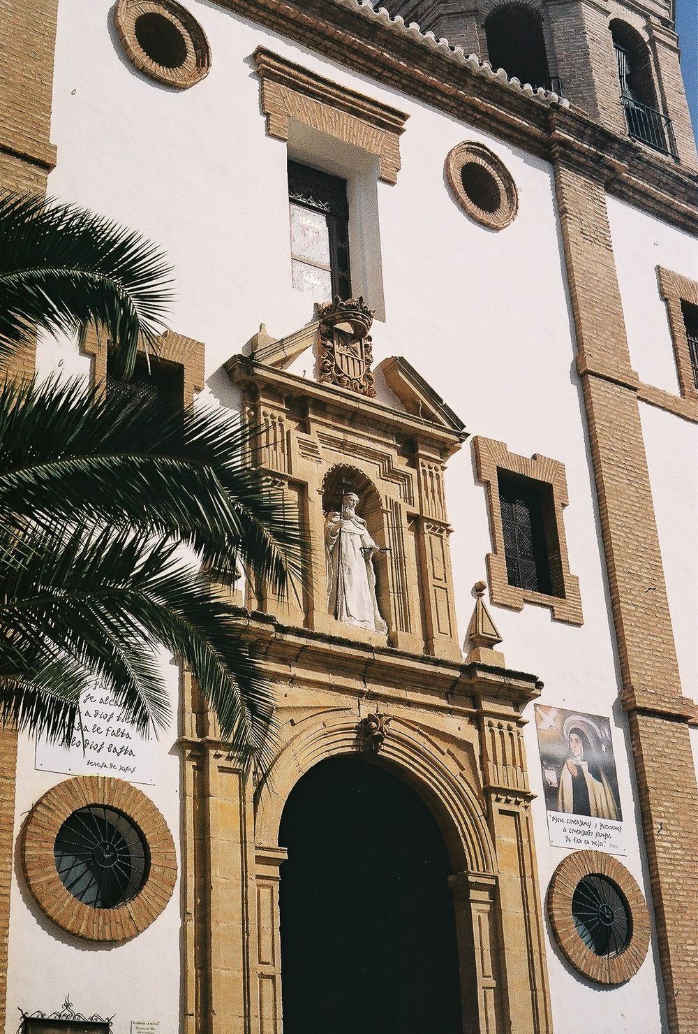 Historic Spanish church facade with ornate baroque portal, religious statue, and palm tree in warm sunlight.