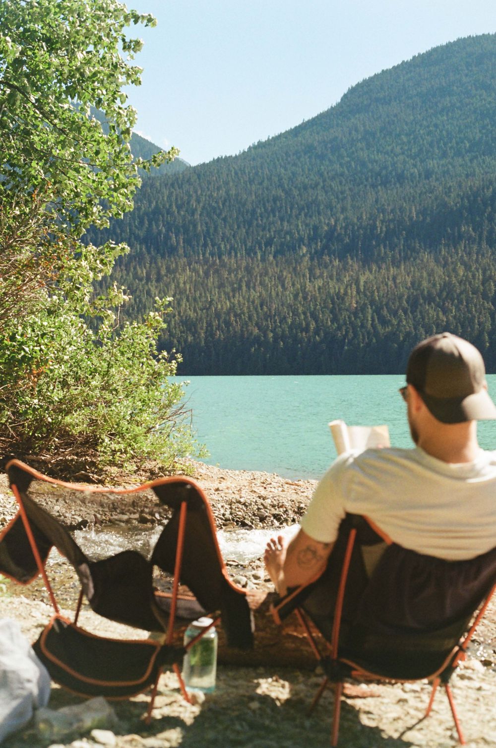 Man reading a book in a camping chair by a turquoise mountain lake surrounded by lush green forest.
