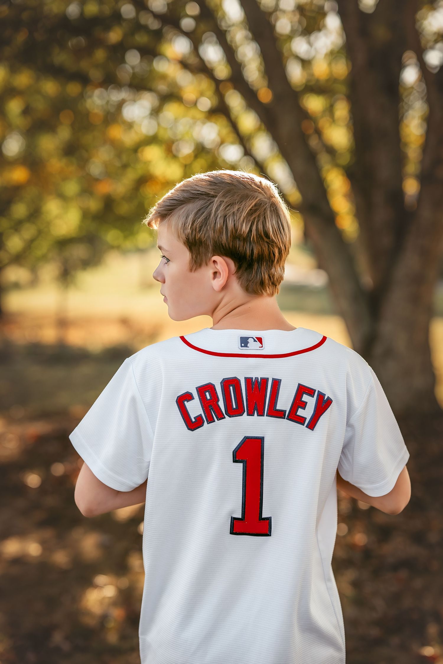 Boy wearing a white Crowley number 1 MLB baseball jersey standing outdoors among autumn trees.