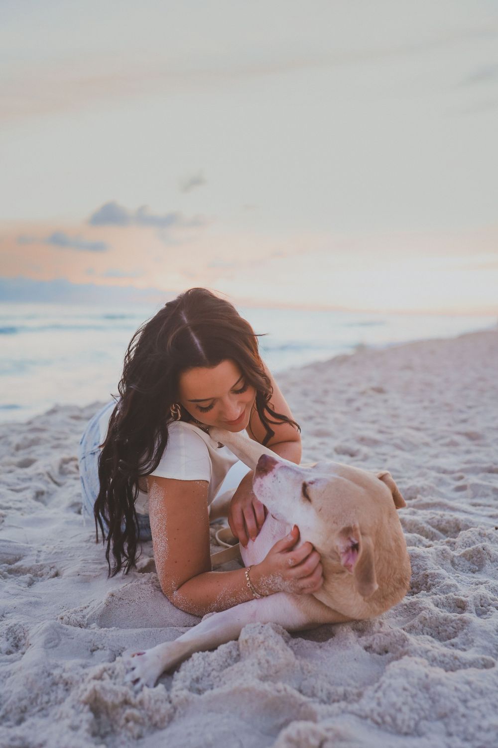 Woman with long dark hair cuddling a yellow Labrador on a sandy beach at sunset.